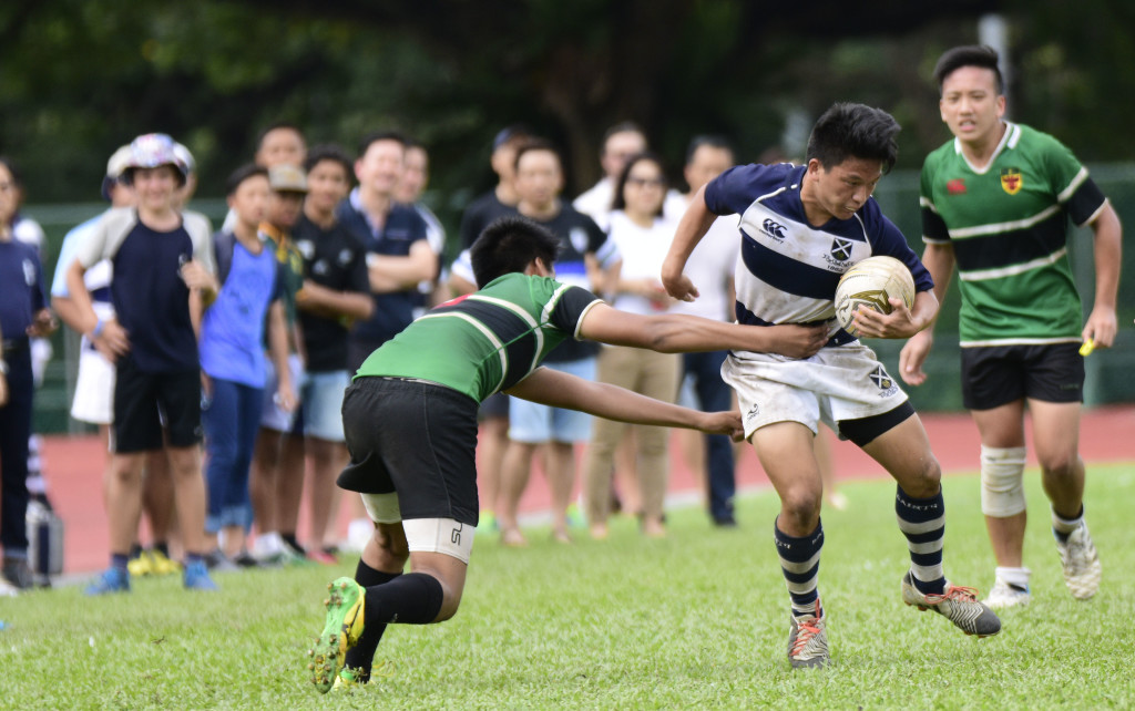 Supporters anxiously looking on as a player from Raffles Institution (left) attempts to tackle his opponent from Saint Andrews (right). (Photo Credit: Eric Lim/ Singapore Cricket Club)